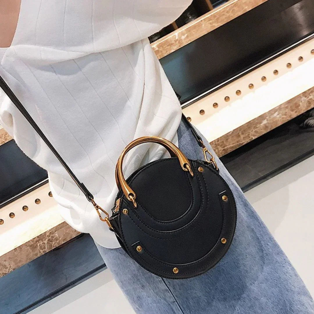 Round black crossbody handbag with gold studs, worn with white blouse and blue jeans