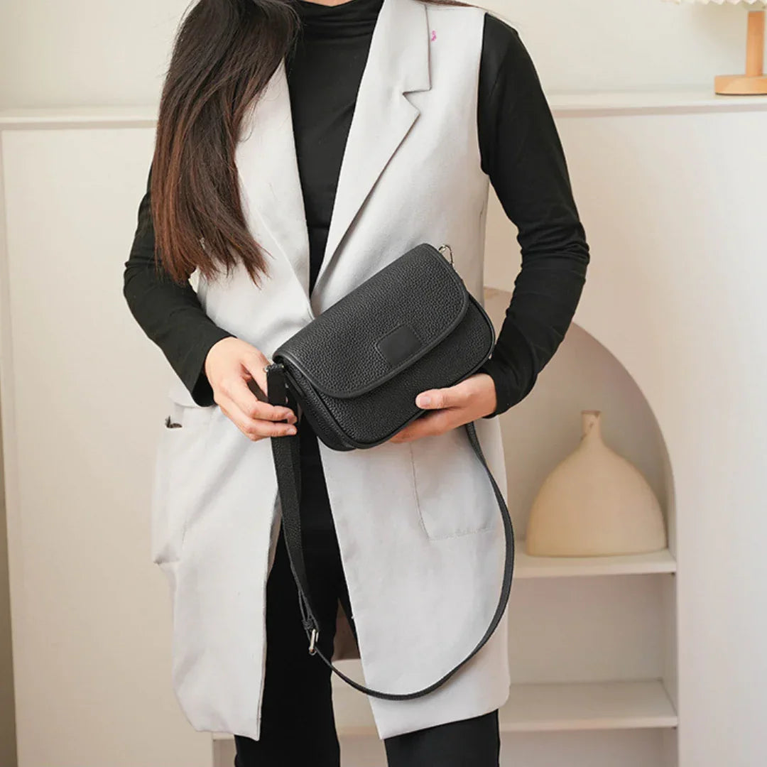 Woman in black top and gray vest holding a black leather crossbody bag indoors