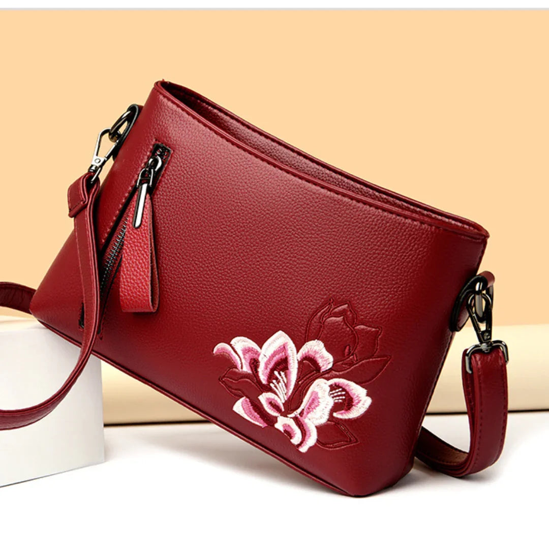 Red leather crossbody bag with pink embroidered flower and zipper, displayed on beige background