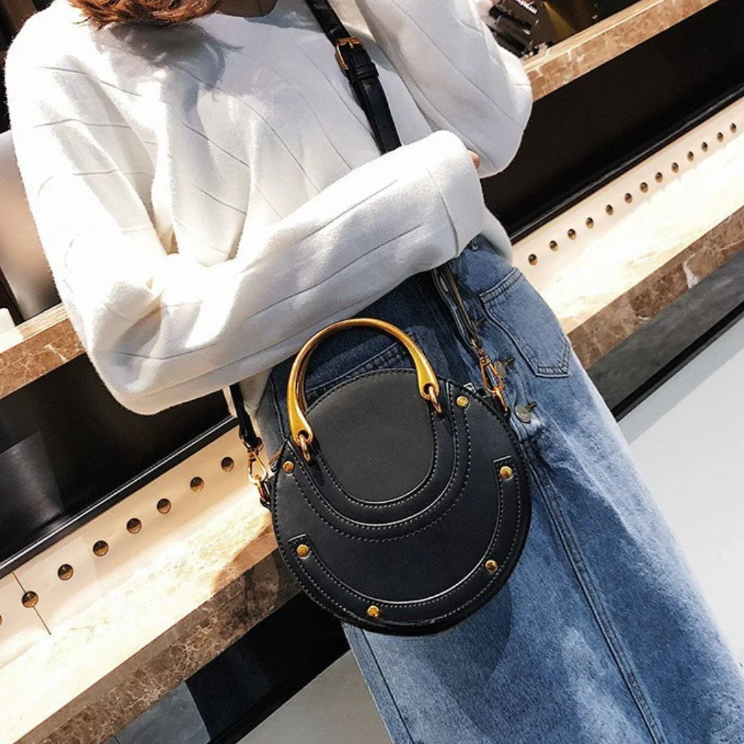 Woman wearing white sweater and denim skirt with round black crossbody purse, gold studs