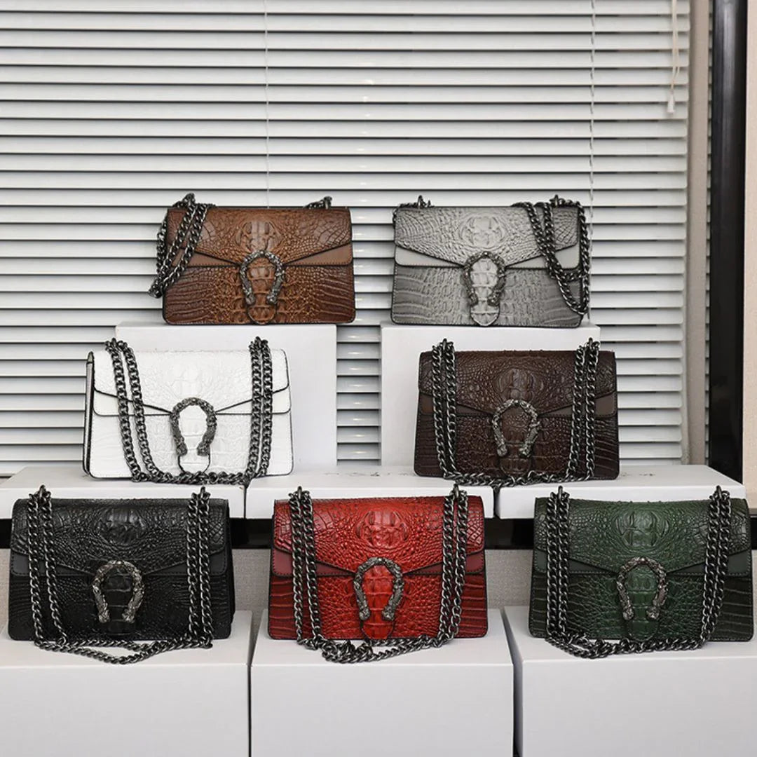 Six crocodile-embossed leather handbags with chain straps in various colors on display boxes.