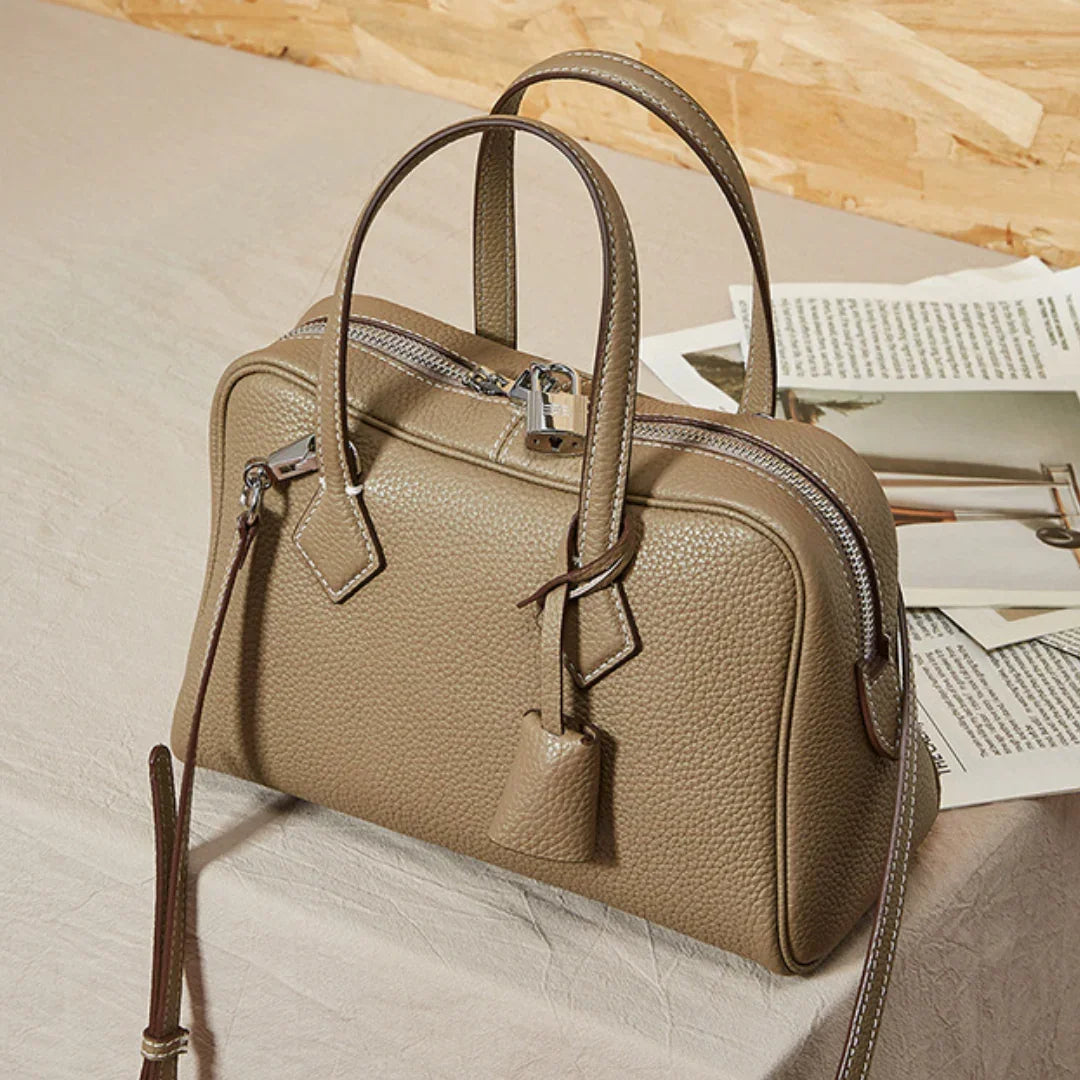 Taupe leather handbag with silver zipper and lock on beige table near open magazines