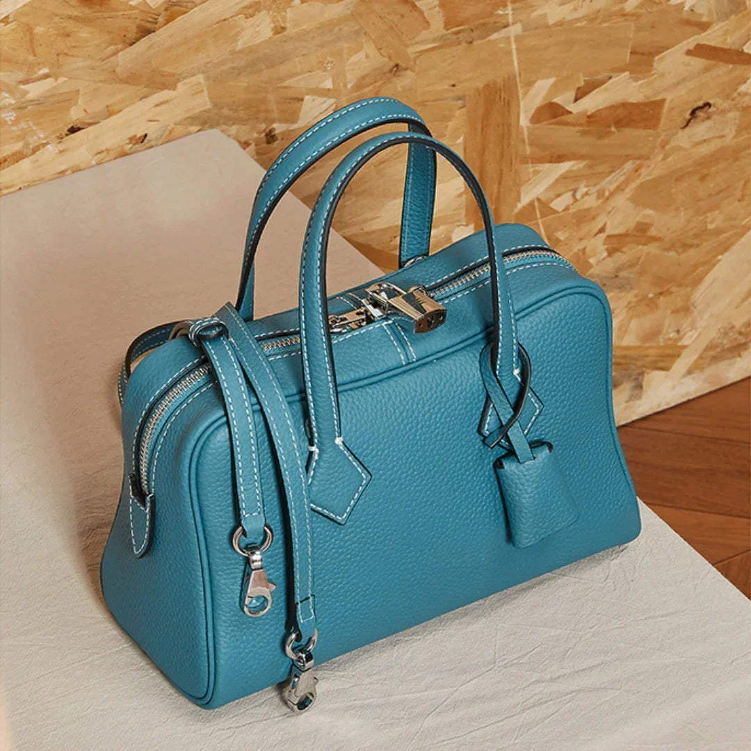 Teal leather handbag with silver hardware and lock on a light surface, wood background
