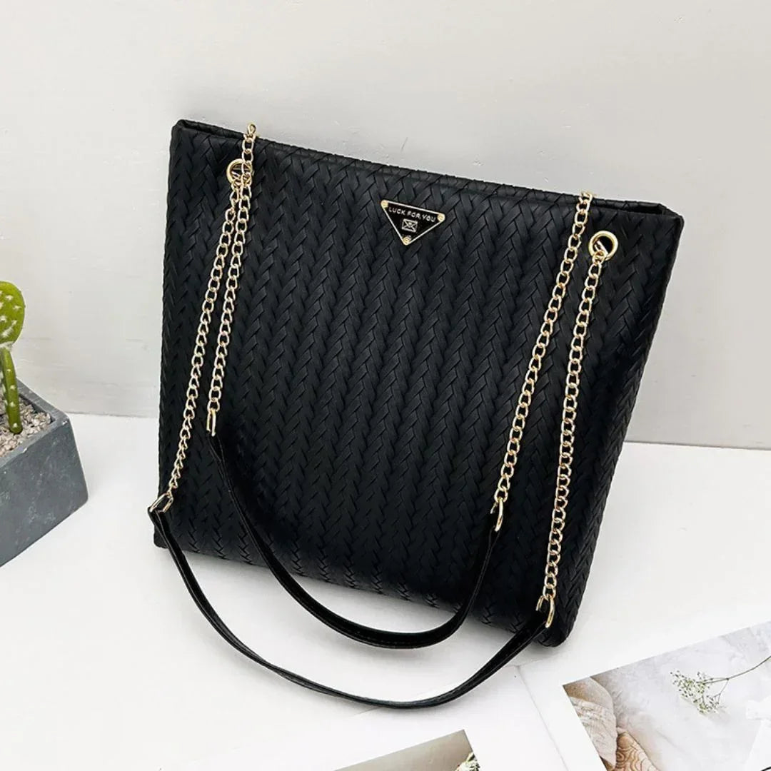 Black woven tote bag with gold chain straps on a white table, modern elegant purse