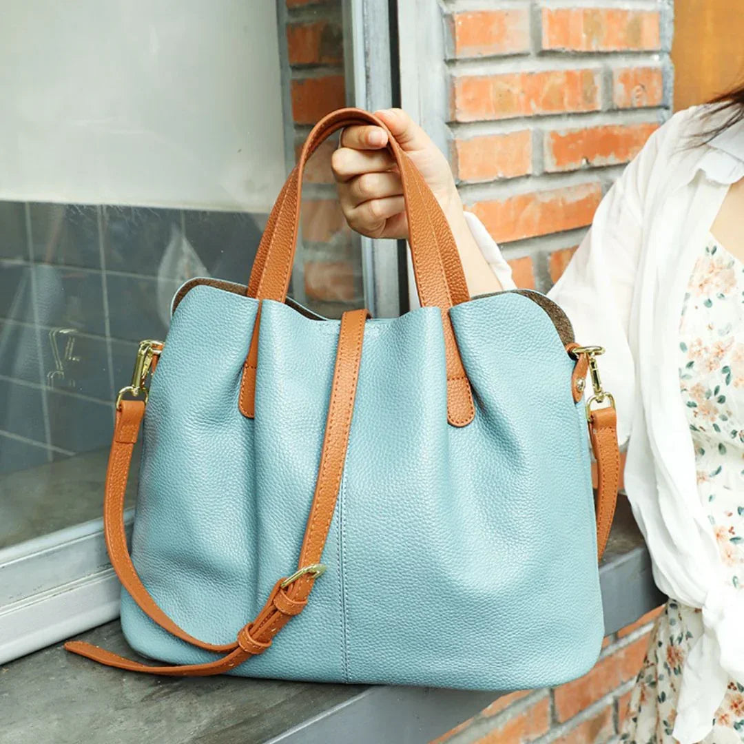 Light blue leather tote bag with brown handles and strap, held by woman near brick wall