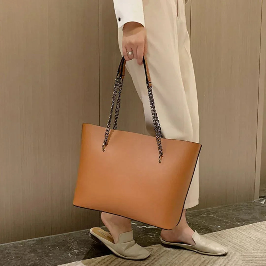 Woman holding brown leather tote bag with chain straps, wearing beige pants and loafers indoors