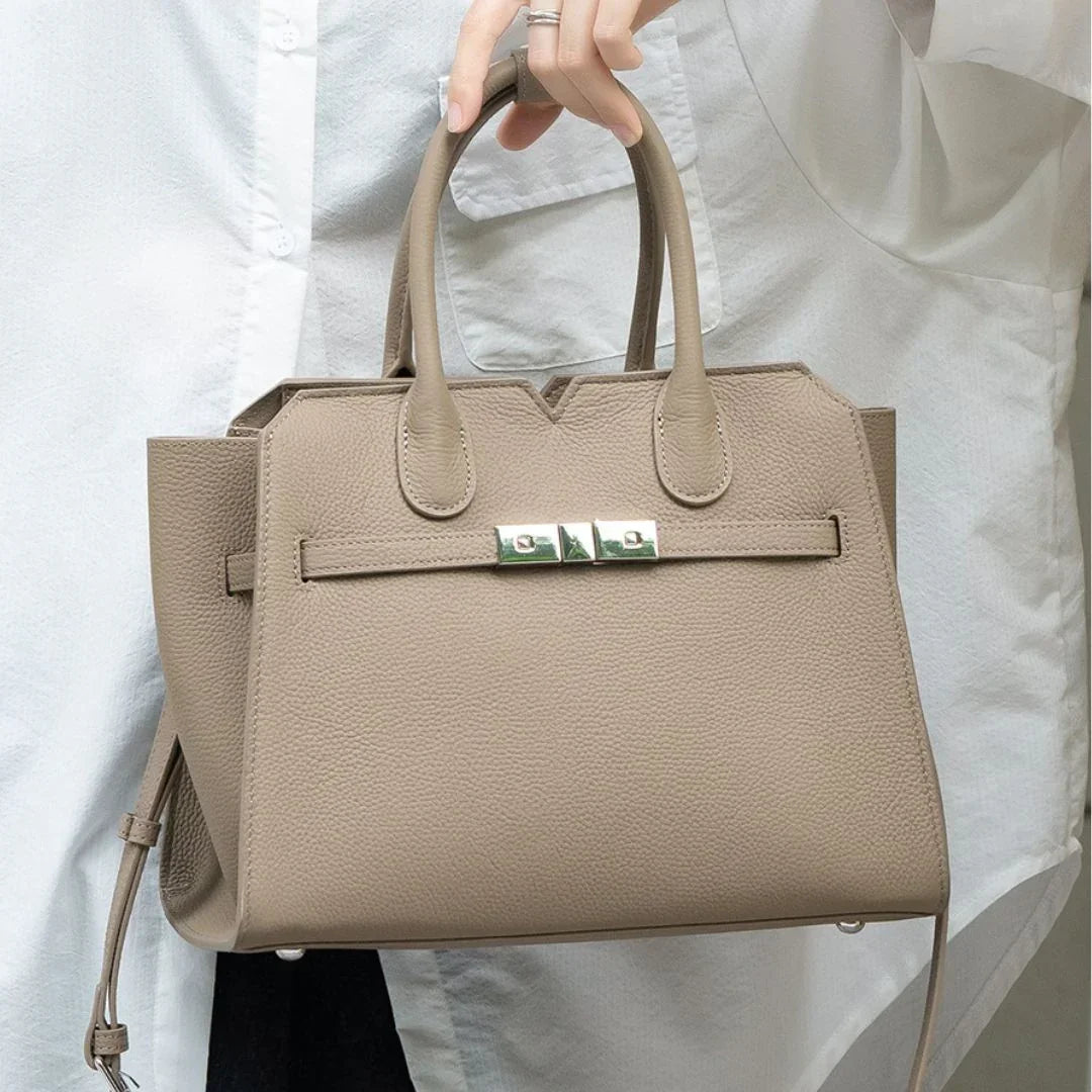 Taupe leather handbag with top handles and silver hardware, held by person in white shirt