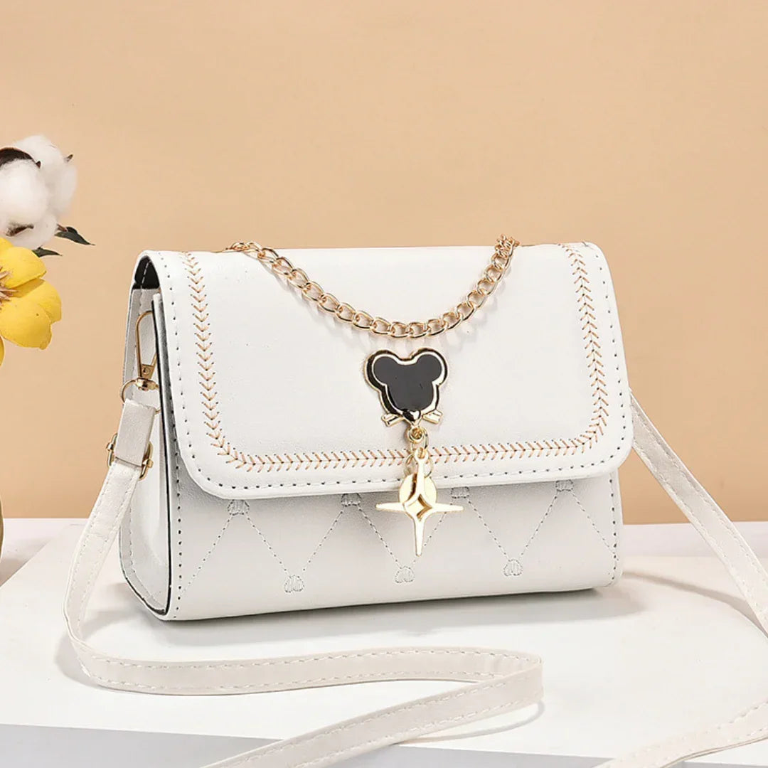 White crossbody bag with gold chain and black bear pendant, on beige background