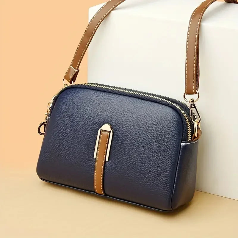 Navy blue leather crossbody bag with brown strap and gold zipper on beige background