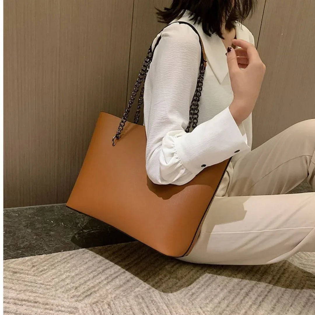 Woman holding large brown leather tote bag with chain strap, wearing white blouse and beige pants