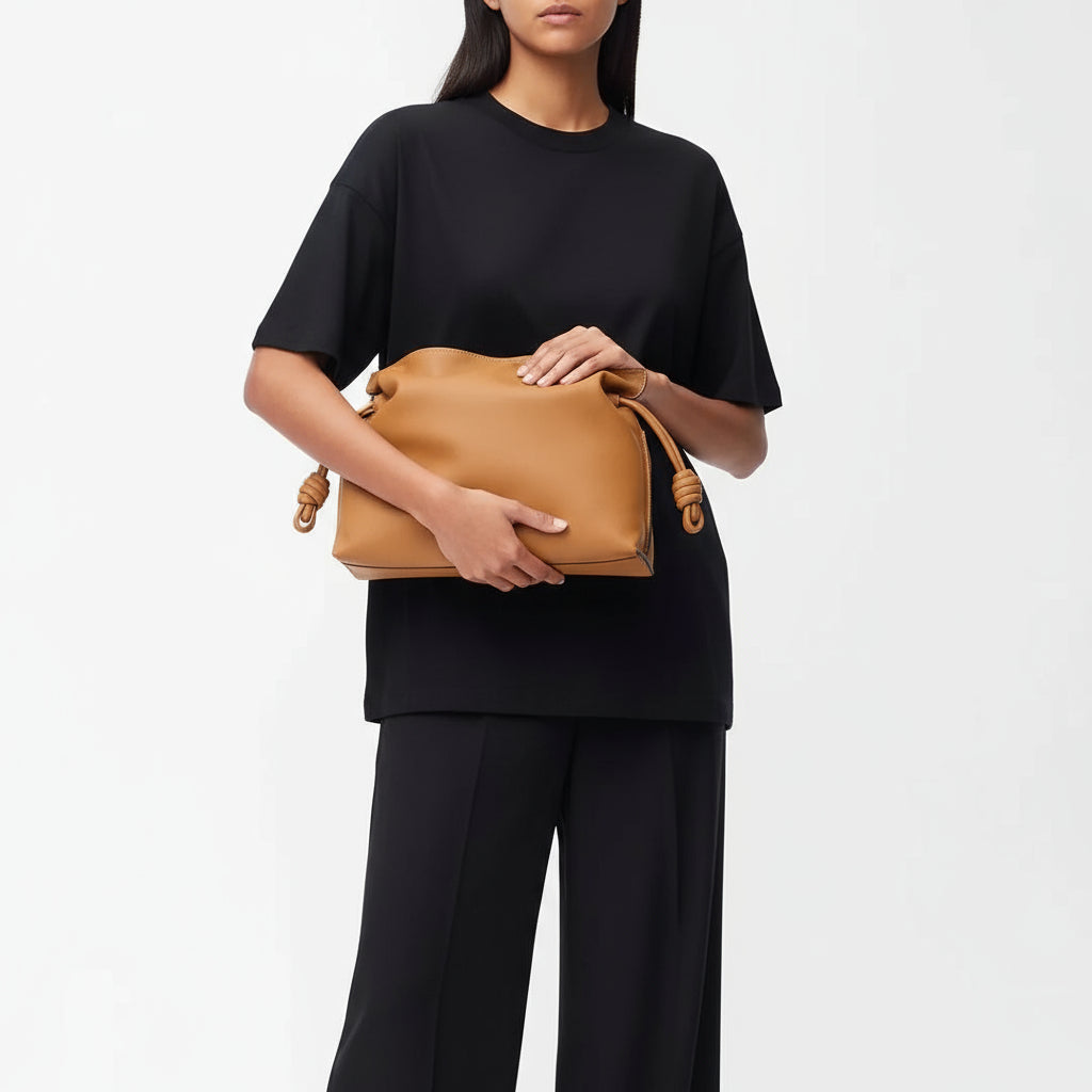 Woman in black outfit holding tan leather handbag with knot detail against plain background