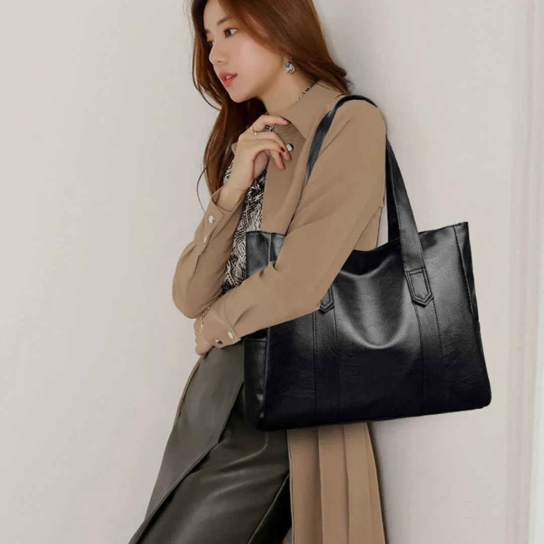 Woman in beige coat carrying a large black leather tote bag against plain wall