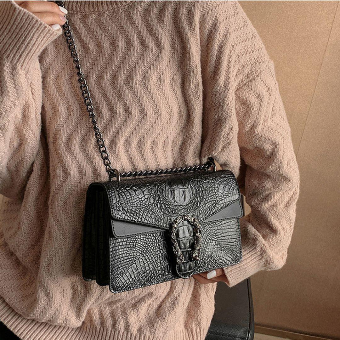 Black crocodile embossed shoulder bag with chain strap and textured clasp, worn over pink sweater