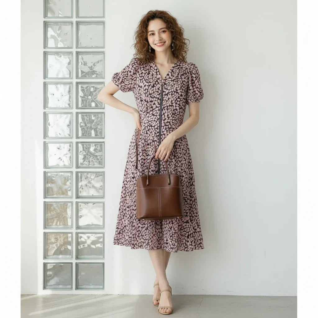 Woman in a purple floral midi dress holding a brown leather handbag, standing by glass blocks