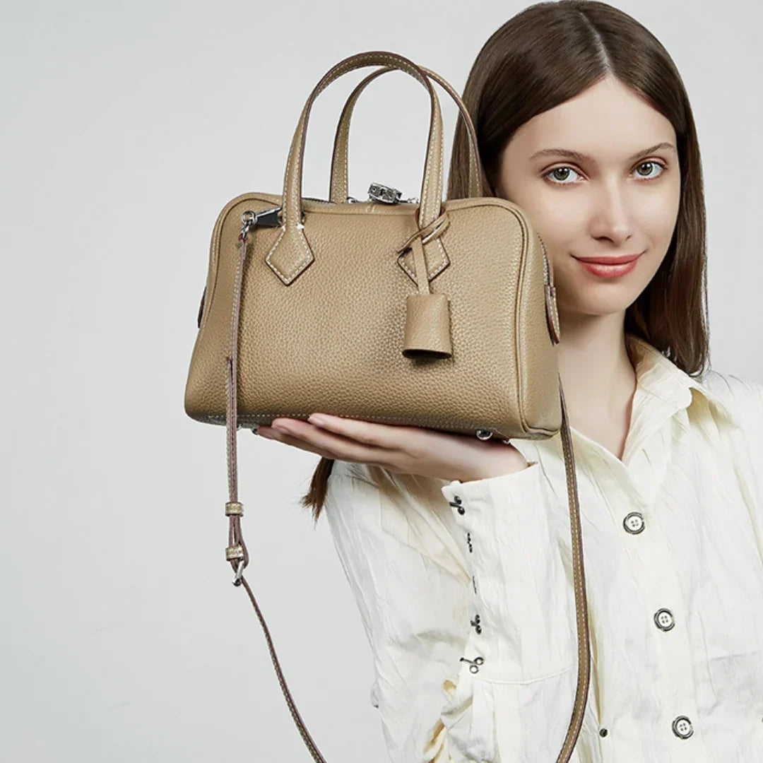 Woman holding a tan leather handbag with top handles and crossbody strap against a plain background