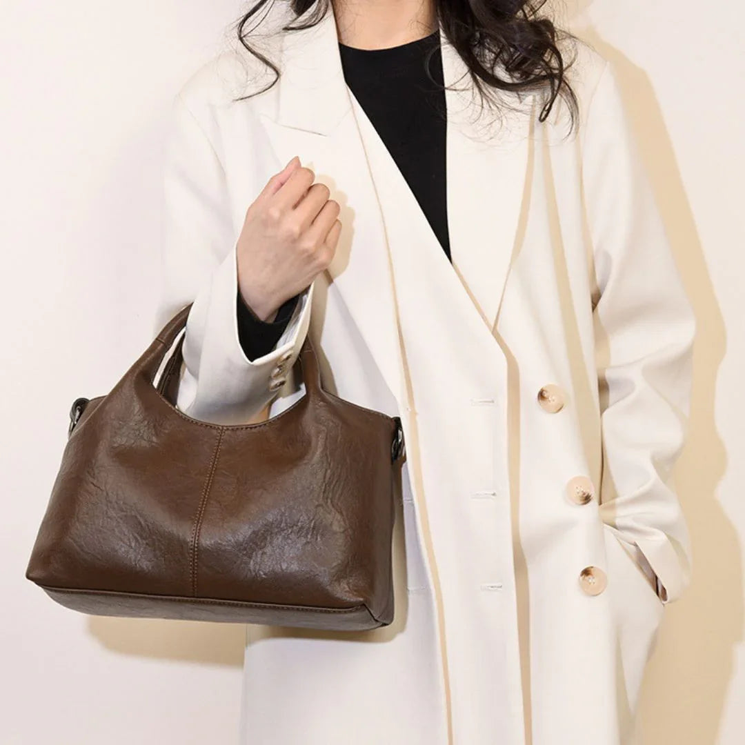 Woman in white coat holding a brown leather handbag against a plain background