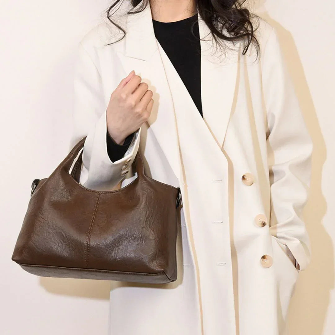 Woman in white coat holding a brown leather handbag against a plain background