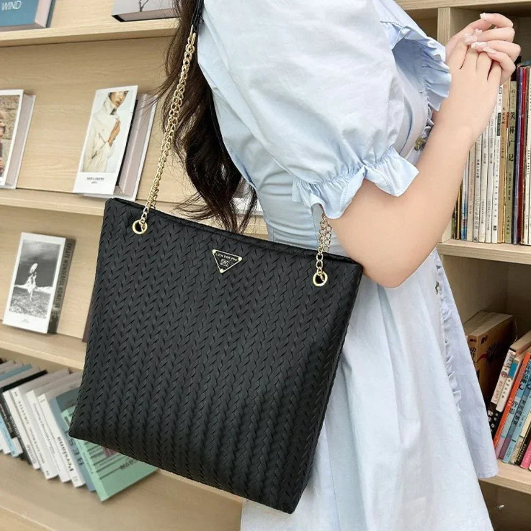 Woman wearing a light blue dress carrying a black woven tote bag with gold chain strap, in a library.