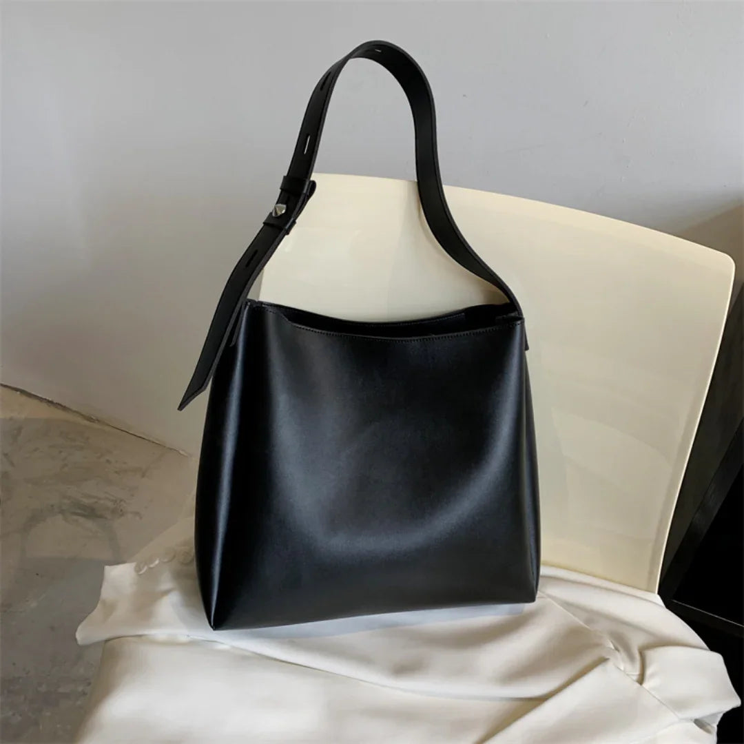Black leather tote bag with adjustable strap on a cream chair