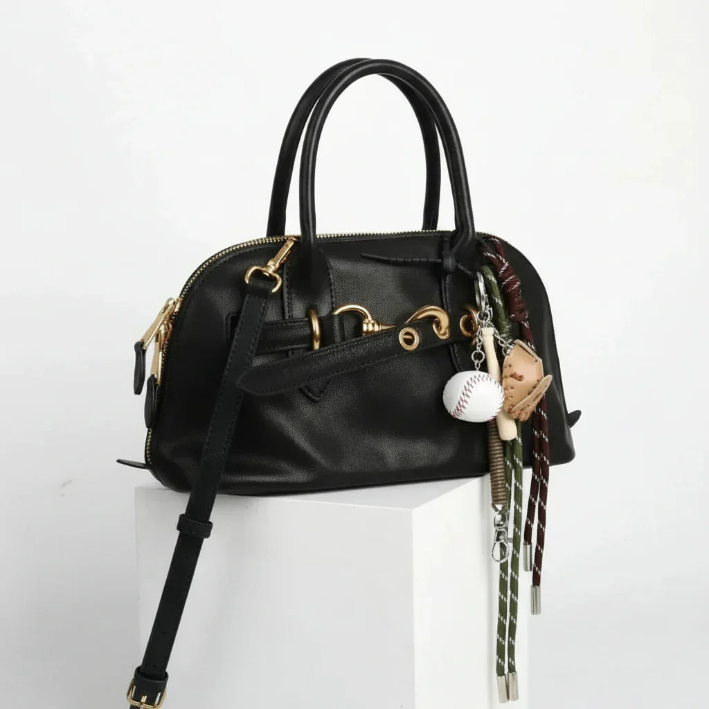 Black leather handbag with gold hardware and baseball-themed keychain, white background