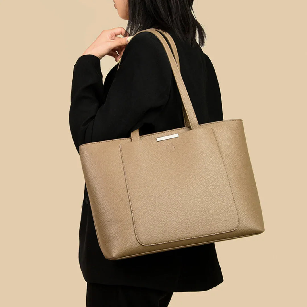 Woman in black blazer carrying a large tan leather tote bag against beige background