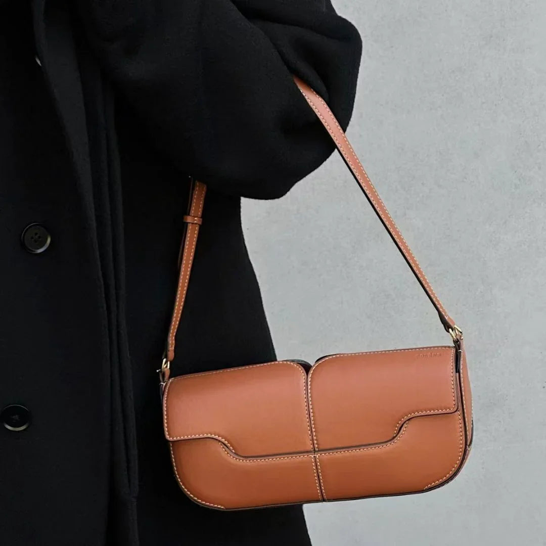 Brown leather shoulder bag with white stitching, worn with a black coat against gray background