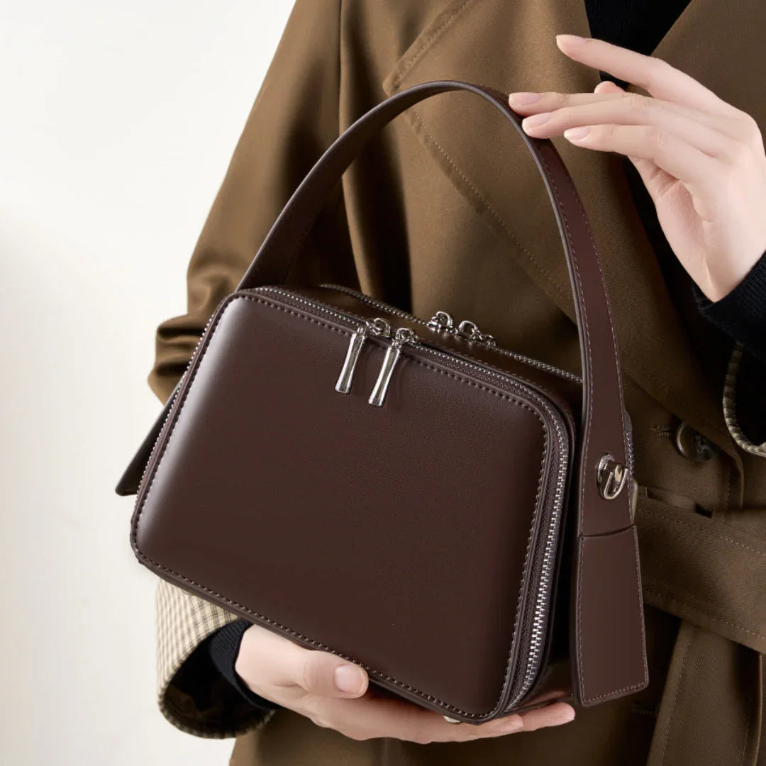 Woman holding brown leather box handbag with silver zippers, wearing brown trench coat