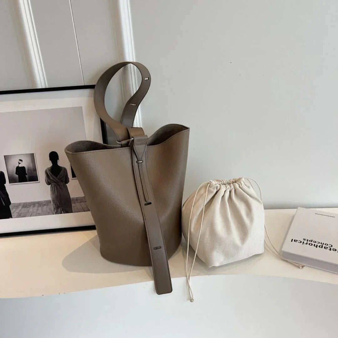 Taupe leather bucket bag and beige drawstring pouch on white table, framed art and book nearby