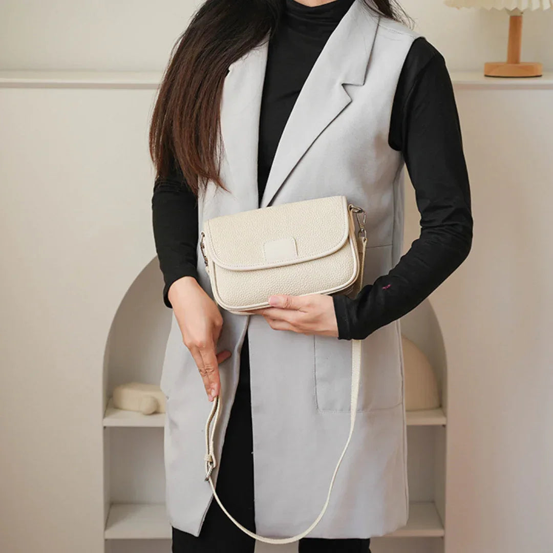 Woman wearing light gray vest and black top holding a beige crossbody bag indoors