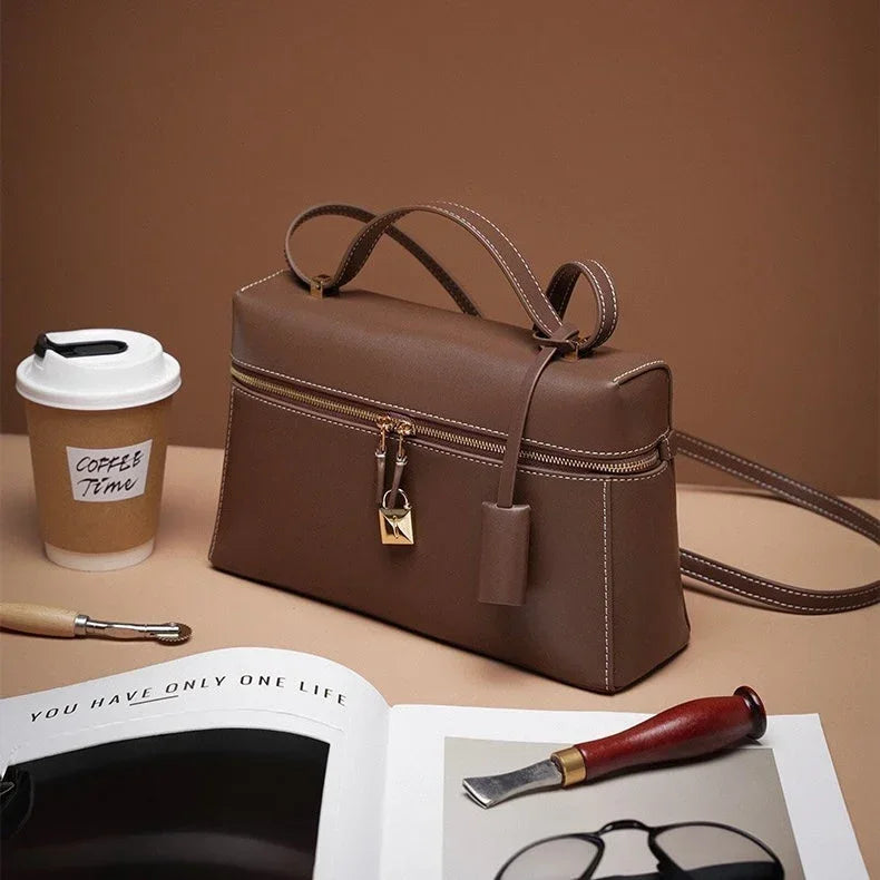 Brown leather handbag with gold zipper and lock, coffee cup, magazine, and carving tool on beige background