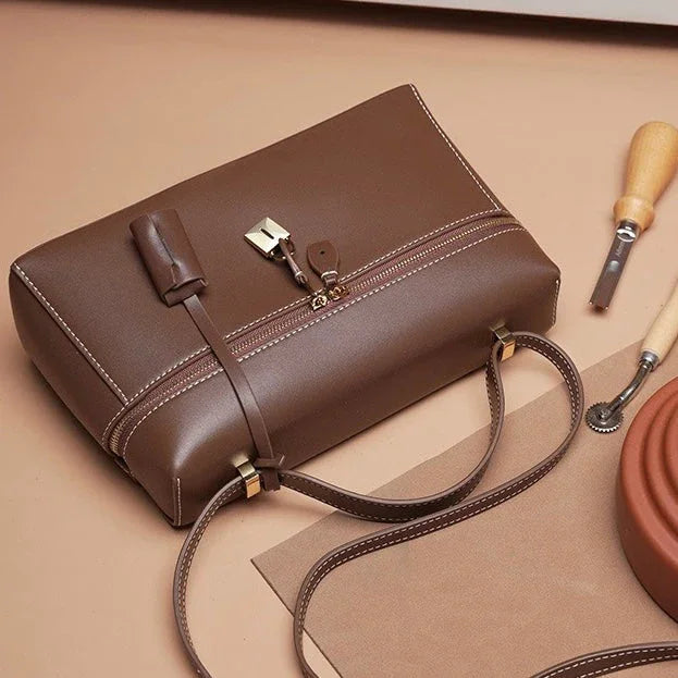 Brown leather handbag with gold hardware on tan background, leathercraft tools beside it