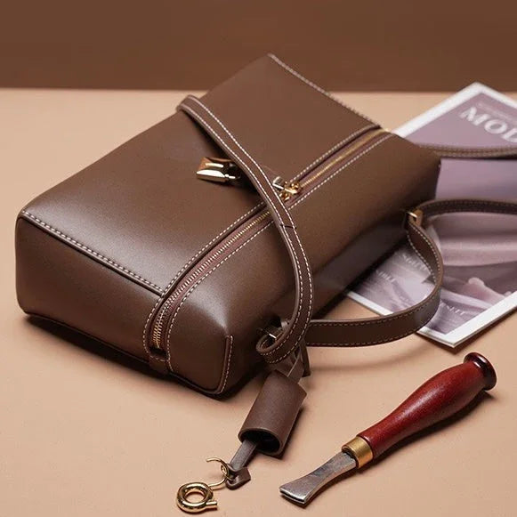 Brown leather crossbody bag with gold zipper, tool, and magazine on tan surface