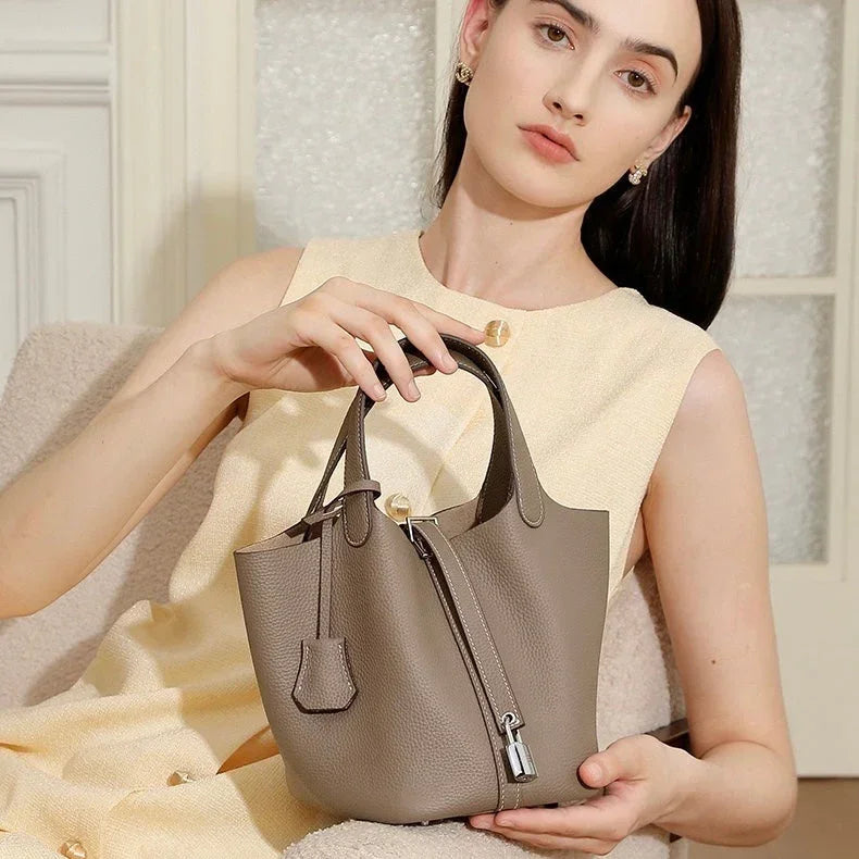 Woman in cream dress holding taupe leather bucket bag with top handles and silver lock