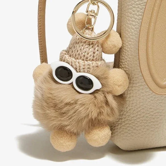 Fuzzy bear pom pom keychain with sunglasses on beige textured handbag
