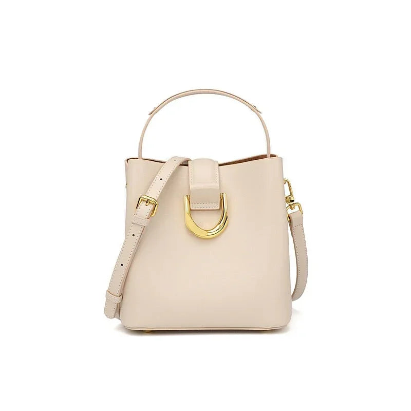 Beige leather handbag with gold buckle, top handle, and crossbody strap on white background