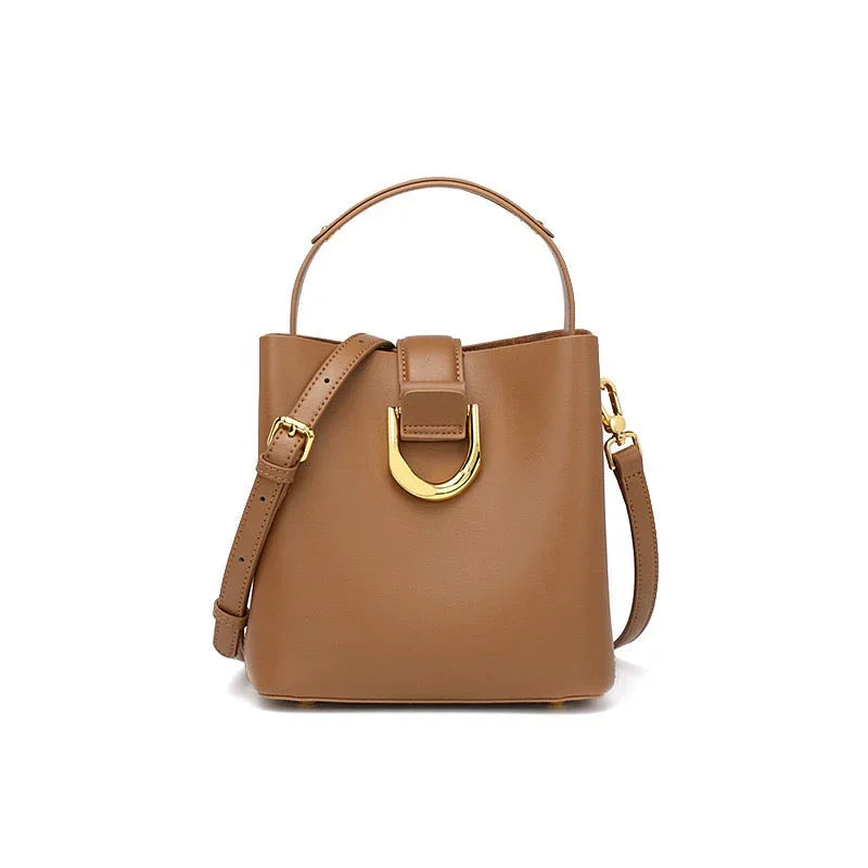 Tan leather bucket bag with gold buckle and adjustable shoulder strap on white background