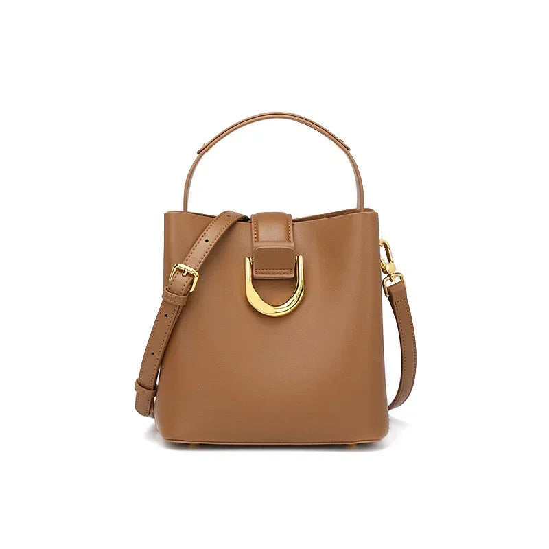 Tan leather bucket bag with gold buckle and adjustable shoulder strap on white background