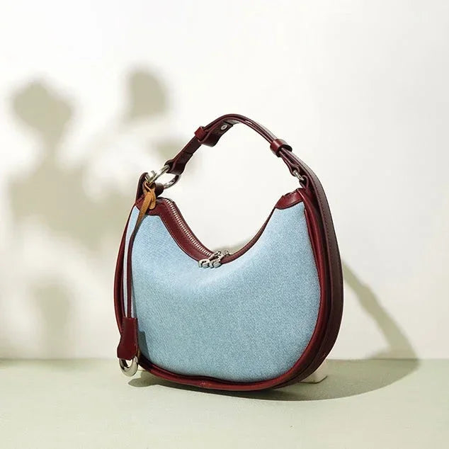 Light blue denim shoulder bag with maroon leather trim and strap on neutral background