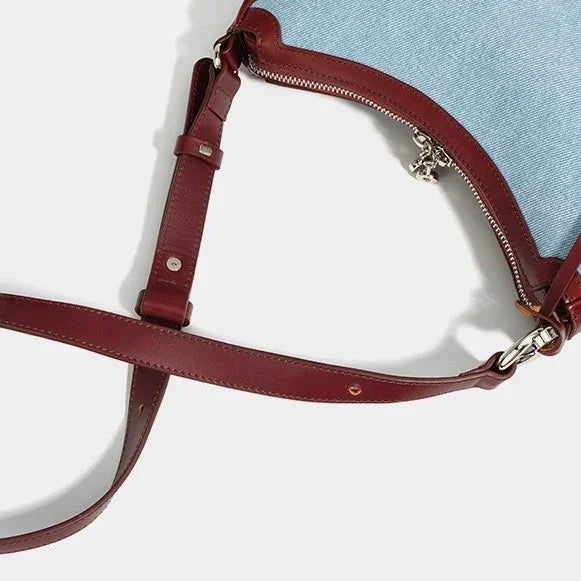 Blue denim handbag with brown leather strap and silver zipper on white background