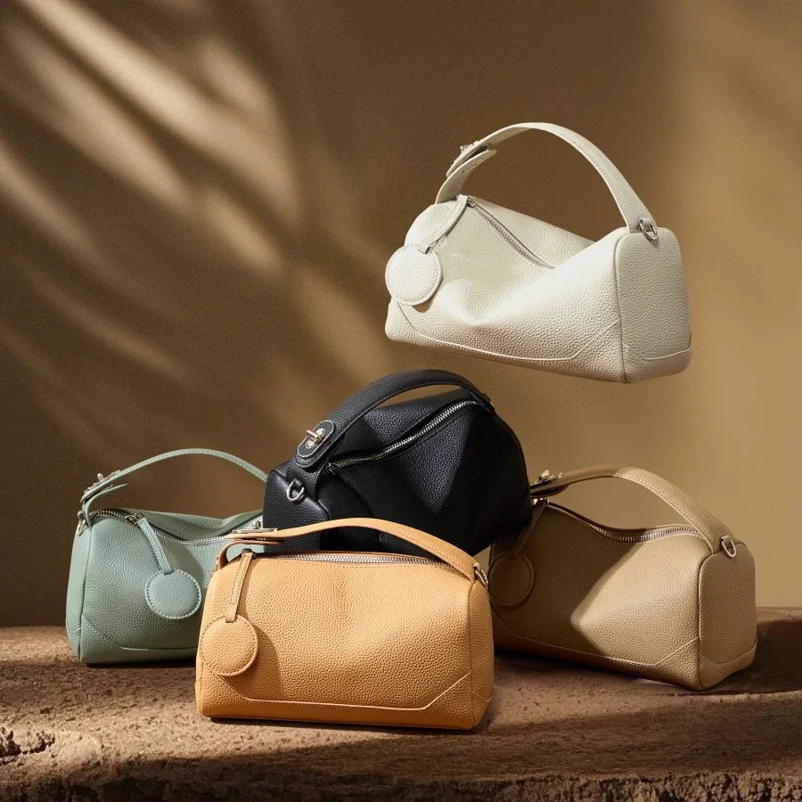 Assorted leather handbags in neutral colors on textured surface with soft brown background