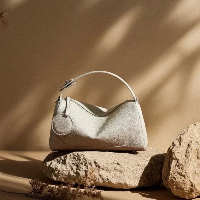 Beige leather handbag with handle, displayed on textured stone, neutral background