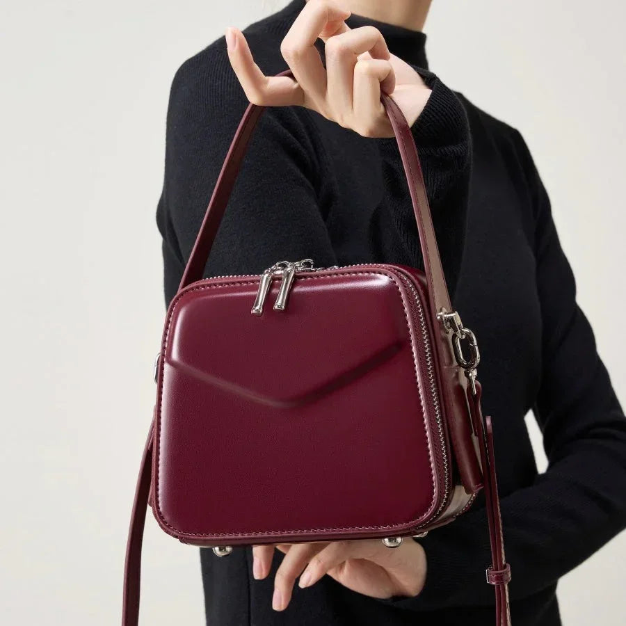 Woman holding a burgundy leather square crossbody bag with silver zippers