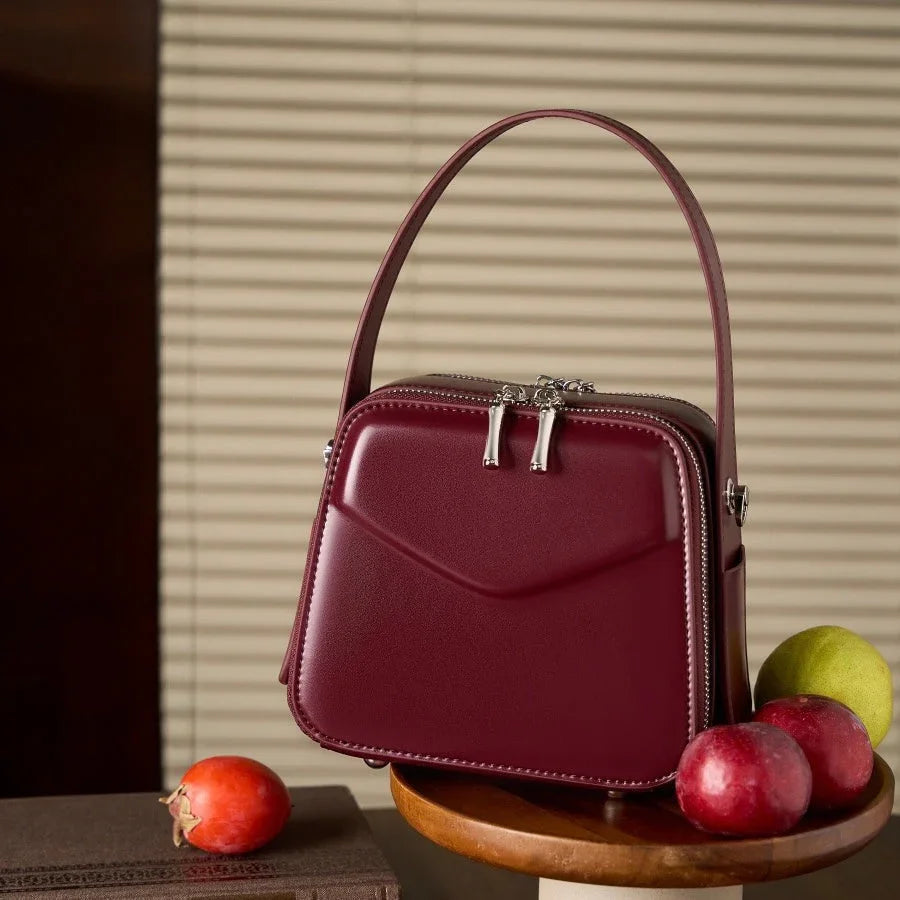 Burgundy leather handbag with silver zippers, styled with apples on a wooden table indoors