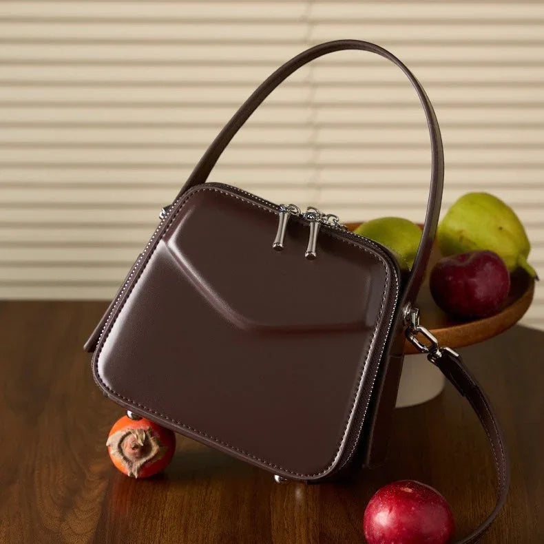 Glossy brown leather handbag on wooden table with fruit bowl and apples in background