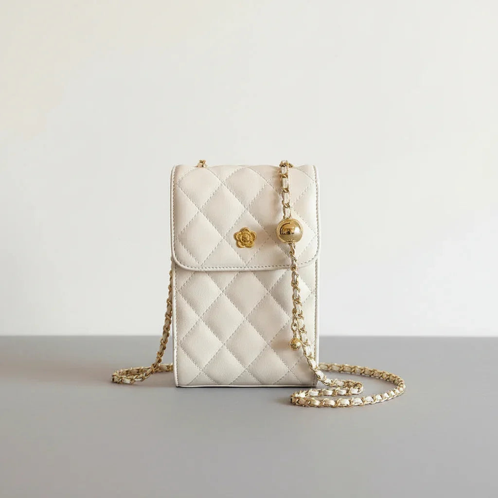 Cream quilted crossbody bag with gold chain strap and flower detail held by hand