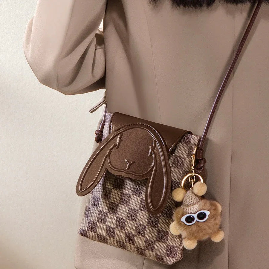 Checkered crossbody bag with bunny face flap and plush keychain, worn over beige coat