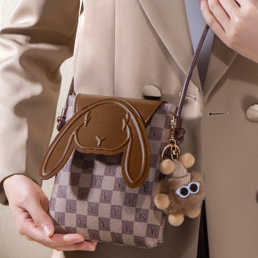 Brown bunny ear crossbody bag with checkered pattern and plush keychain, held by woman in tan blazer