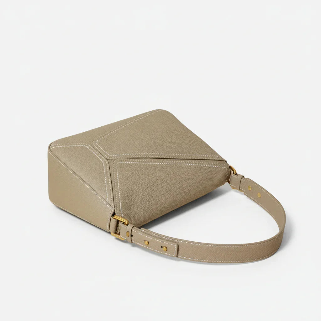 Taupe leather crossbody bag with gold hardware and white stitching on a beige surface