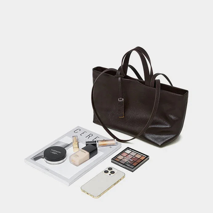Black leather tote bag with cosmetics, iPhone, magazine, and makeup palette on white background