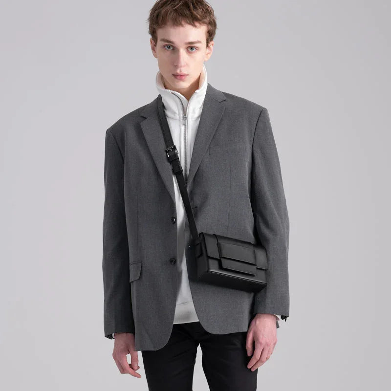 Man in a gray blazer and white zip-up with black crossbody bag, minimalist background